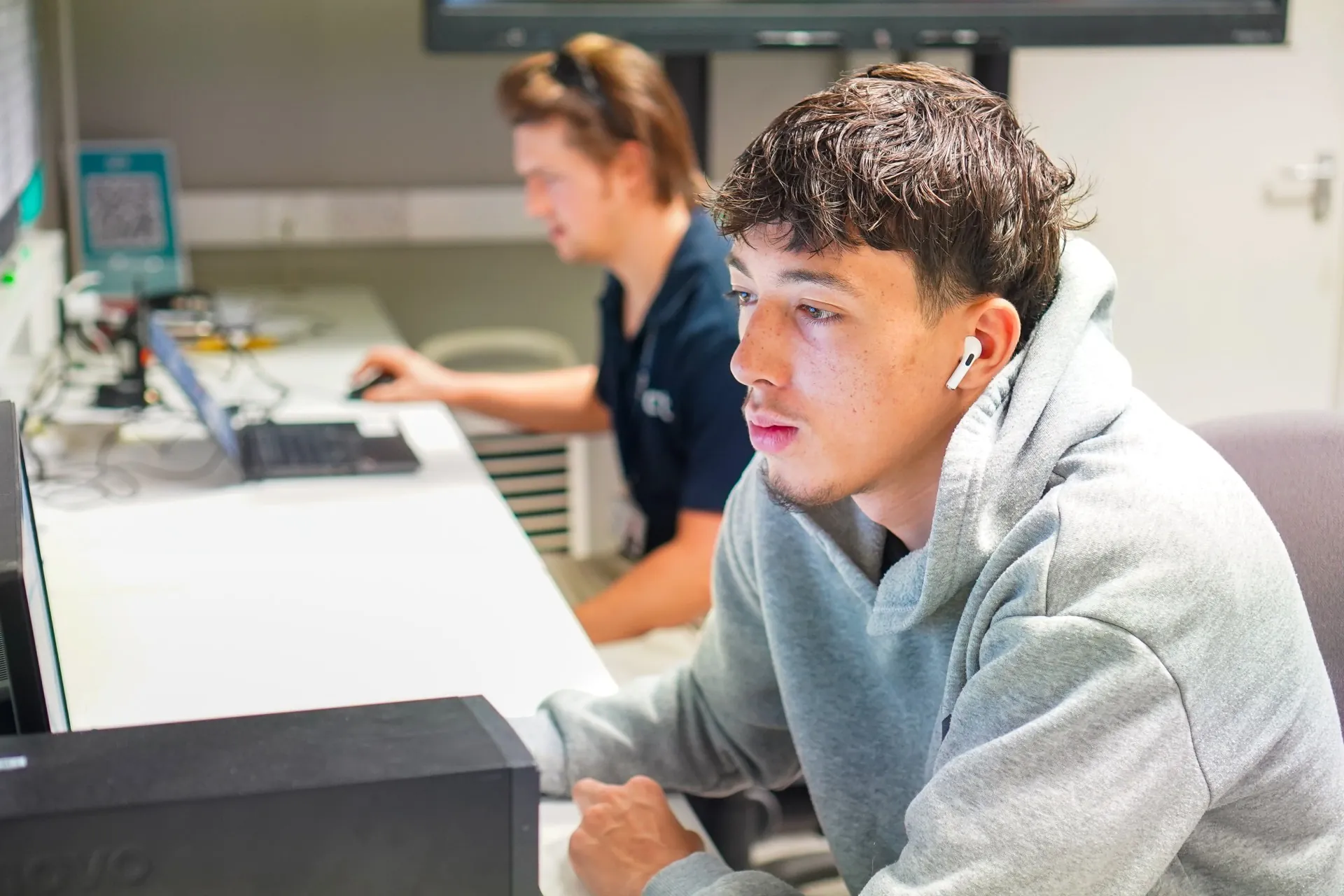 young dark haired learner sat looking at a computer in his classroom. wearing a grey hoodie with act tutor visible in the background